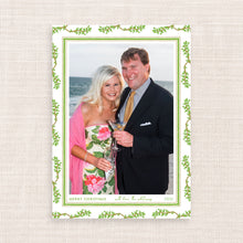 A custom holiday photo card featuring a printed border of green leaves and vines on a white background, with a central photograph of a smiling couple holding drinks. The bottom of the card reads 'MERRY CHRISTMAS with love, the whitmers 2026'.